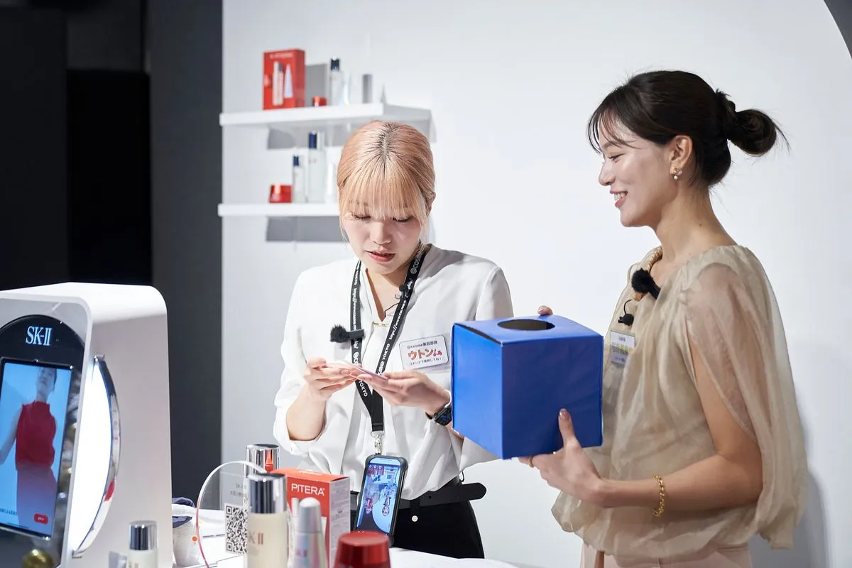 Two hosts at the SK-II booth, one filming a product on her phone while the other holds a blue box.