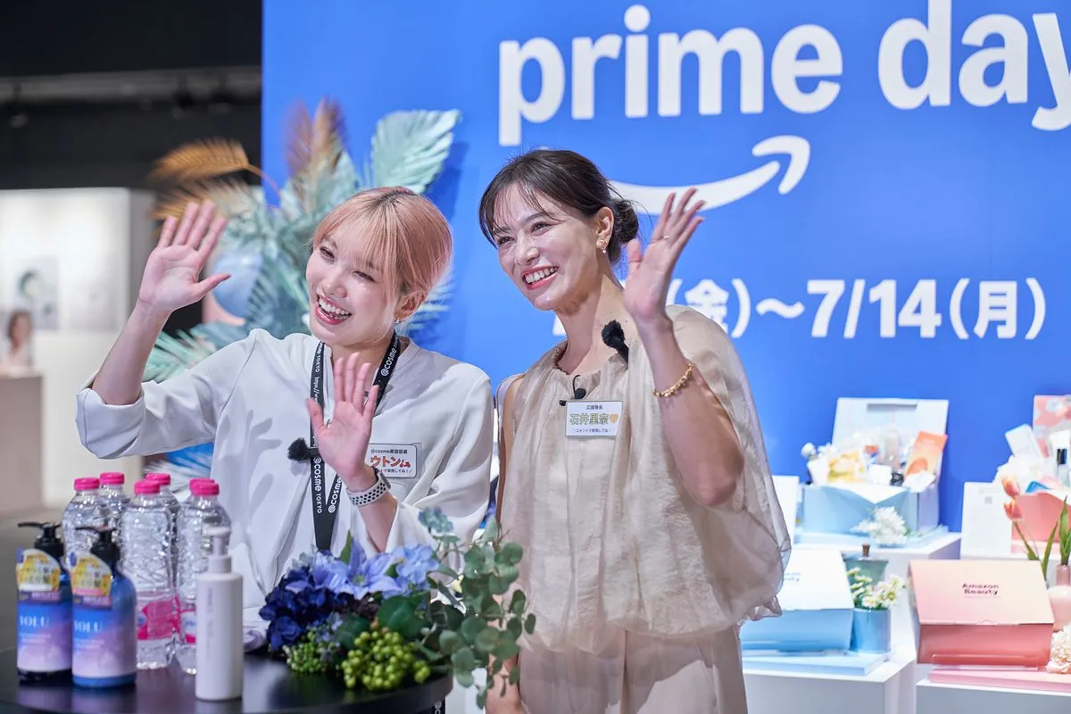 Two hosts wave to the camera at the Amazon Beauty Festival, in front of a blue Prime Day wall with the dates 7/11 to 7/14 and a table of products.