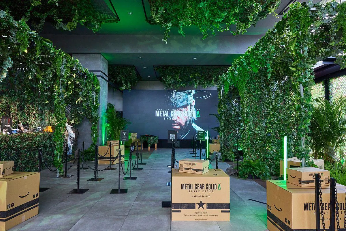 Metal Gear Solid Delta launch event: a walkway lined with hanging ivy and green lights, stacked branded Amazon boxes, and Snake on a large screen.